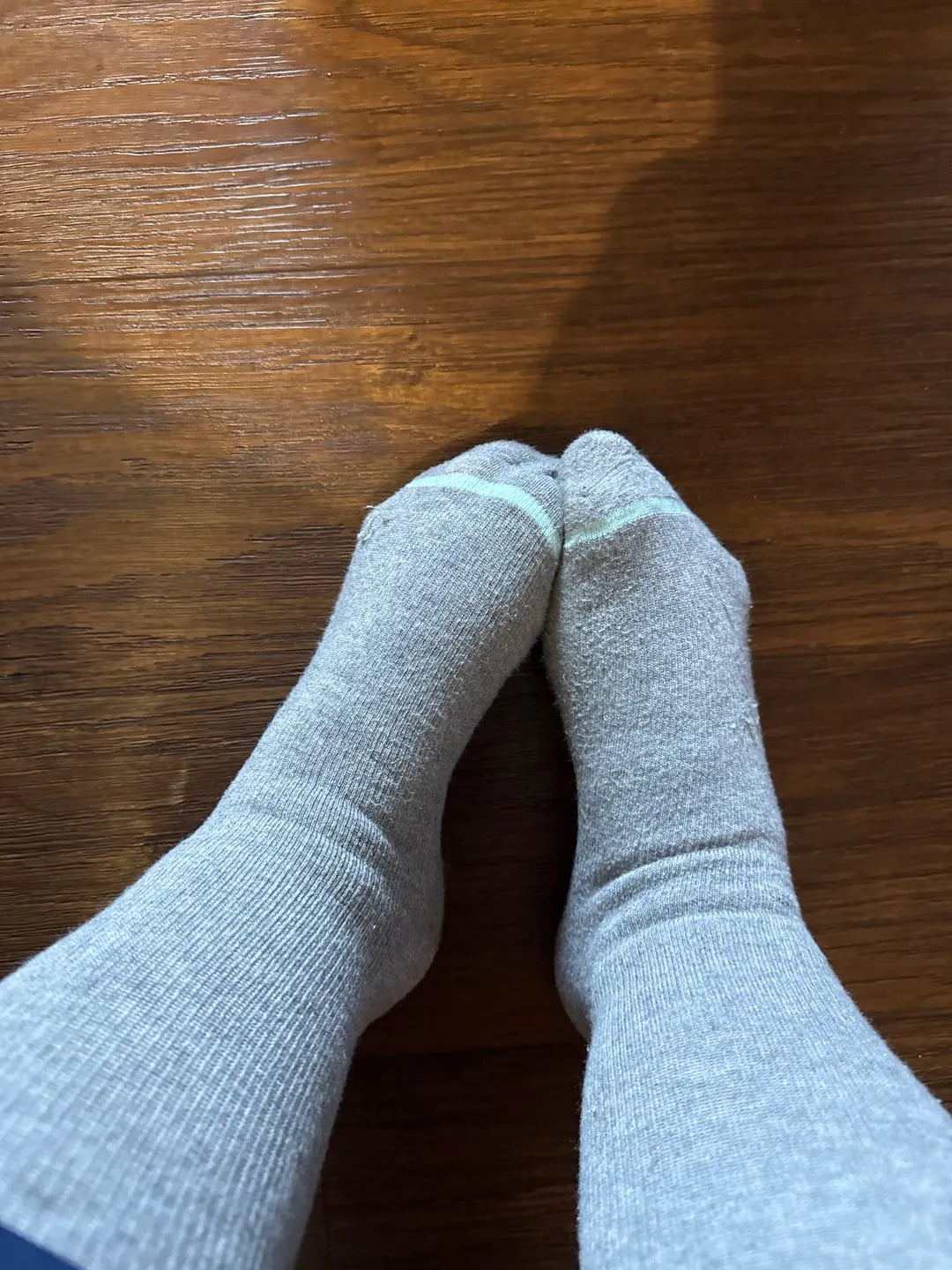 Top-down view of a person's feet wearing light grey bamboo compression socks with a teal toe stripe on a dark wood floor.
