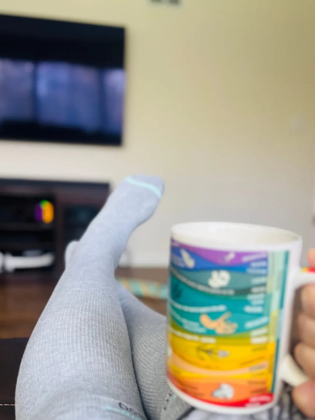 First-person perspective of legs crossed wearing grey bamboo compression socks while holding a colorful mug in a cozy, blurred living room setting.