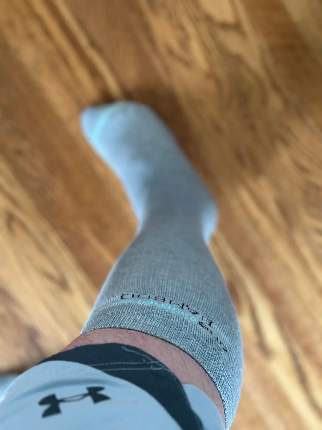 Close-up POV shot of a man wearing a grey bamboo compression sock, showing the calf fit and ribbed texture on a hardwood floor.